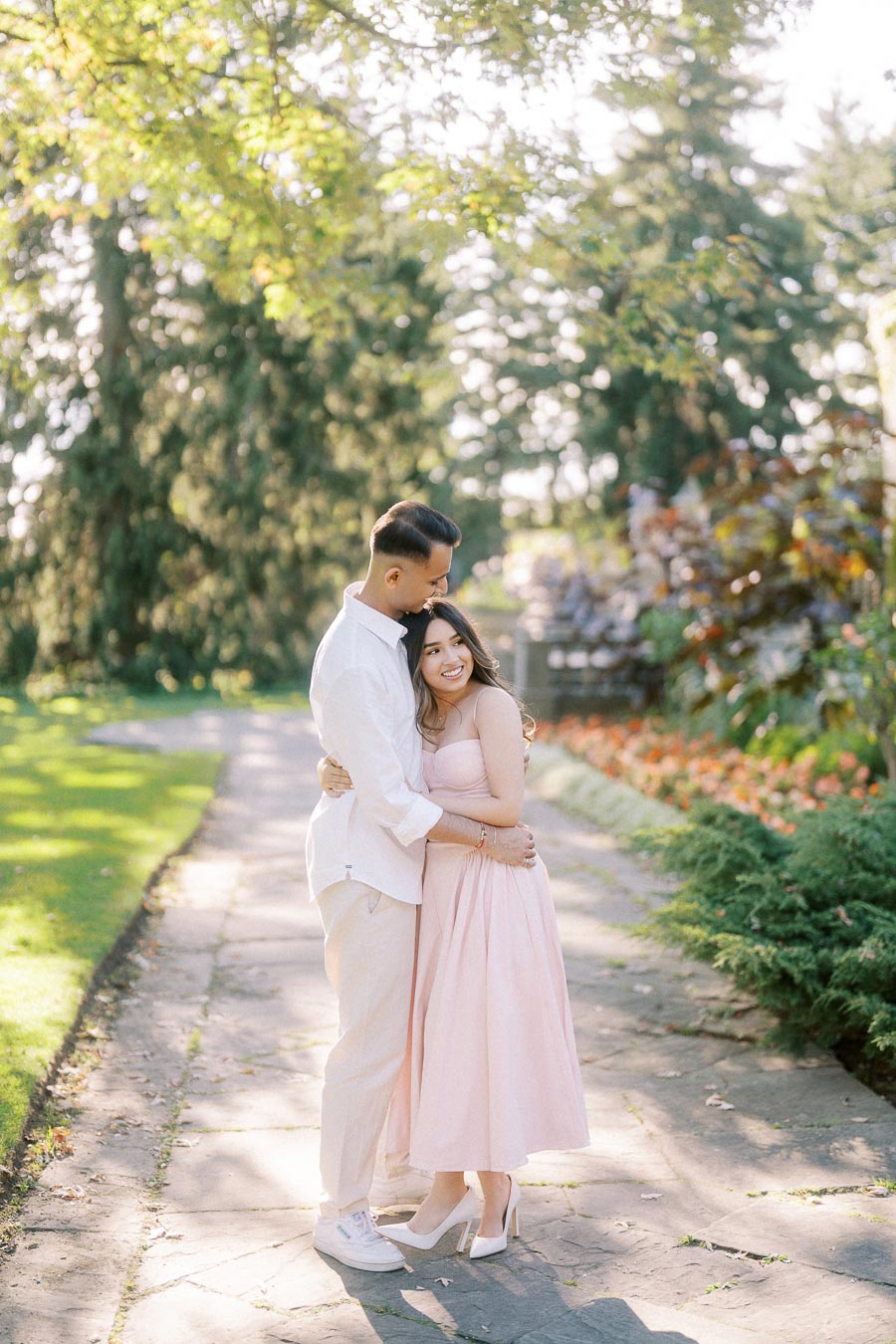 A couple embraces on a sunlit garden pathway, surrounded by lush greenery and blooming flowers, capturing a romantic moment together.