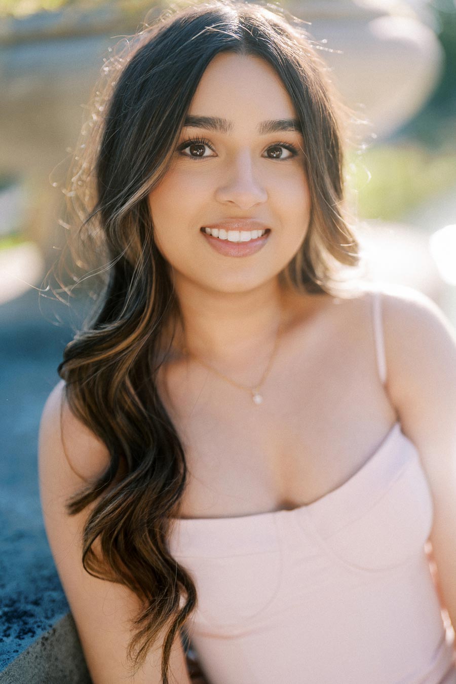Smiling woman with long hair wearing a light-colored camisole in a sunny outdoor setting.