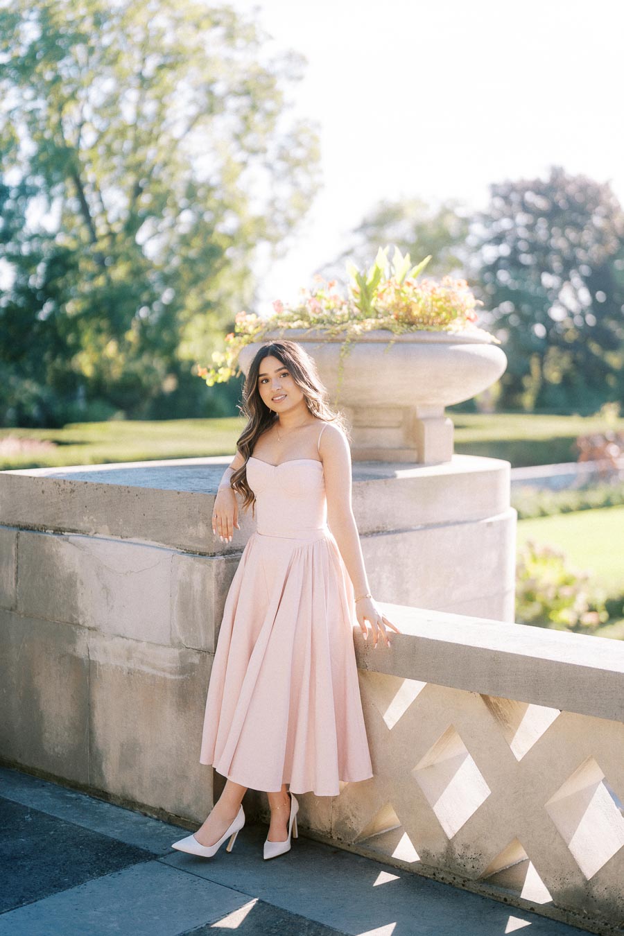 Woman in a blush pink dress standing beside a large stone planter in a sunlit garden setting.