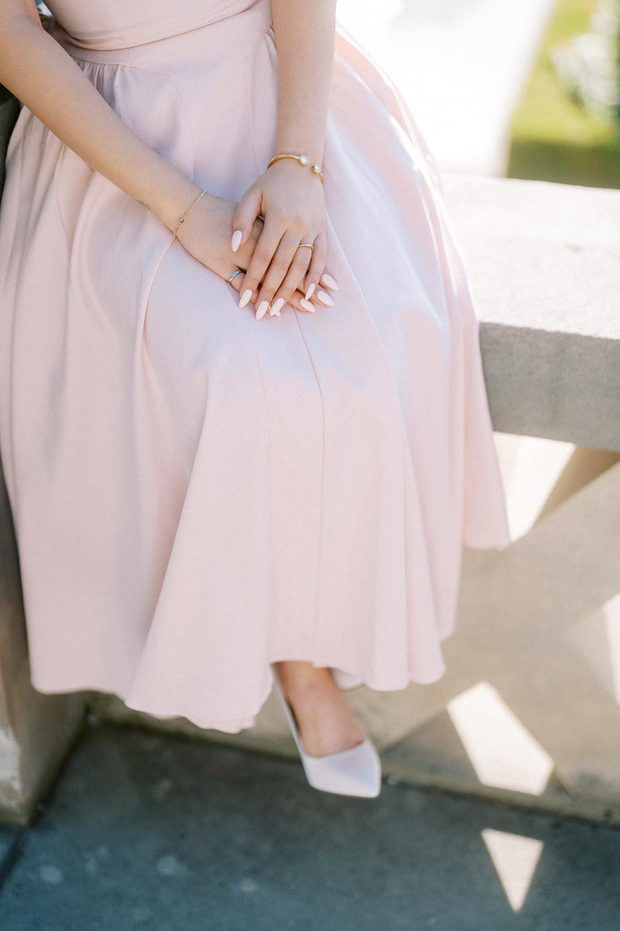 Elegant woman in a light pink dress sitting on a stone bench, showcasing manicured hands with gold bracelets and wearing white pointed-toe heels.