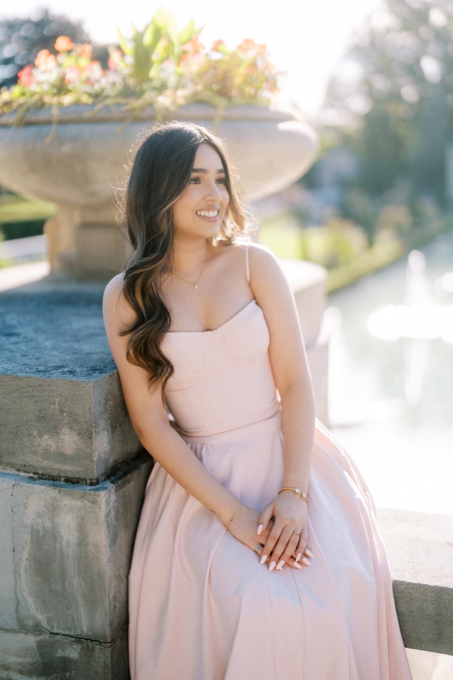 A woman in a light pink dress sits on a stone ledge near a garden planter filled with colorful flowers, enjoying a sunny day.