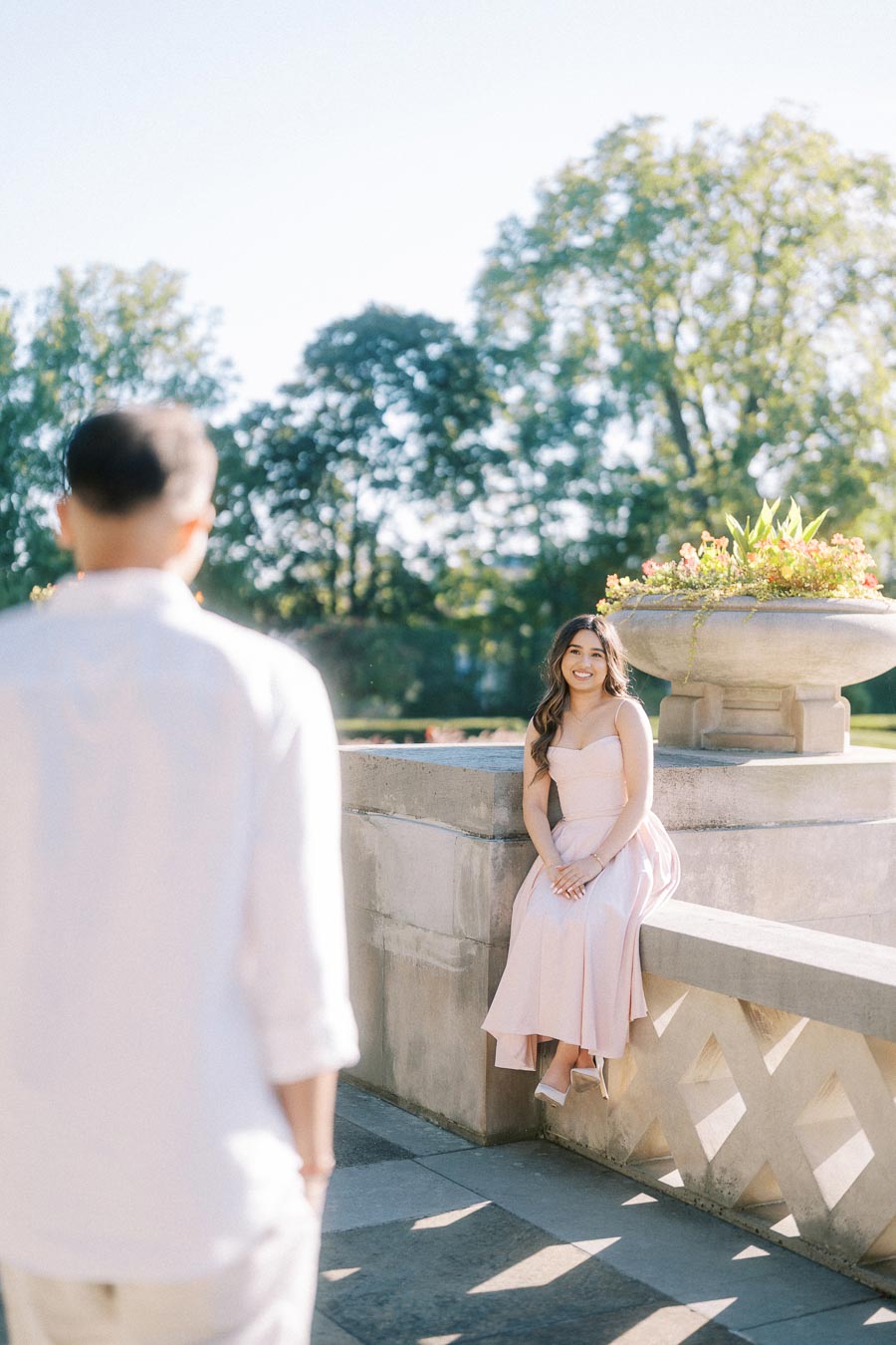 Young woman in a pink dress sitting on a stone railing, smiling towards a person in a white shirt, with lush greenery in the background on a sunny day.