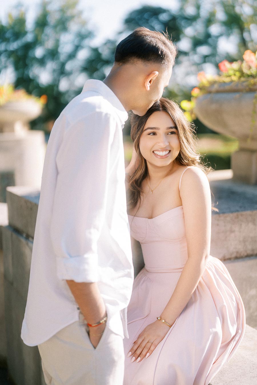 A young couple enjoys a romantic moment outdoors, with the man leaning towards the woman who is seated on a stone ledge, smiling brightly. She is wearing a light pink dress, and the background features blurred greenery and flowers, evoking a warm, sunny day.