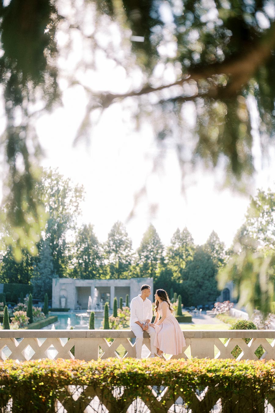 A couple sitting on a stone railing in a sunlit, beautifully landscaped garden with fountains and greenery in the background, framed by soft focus tree branches.