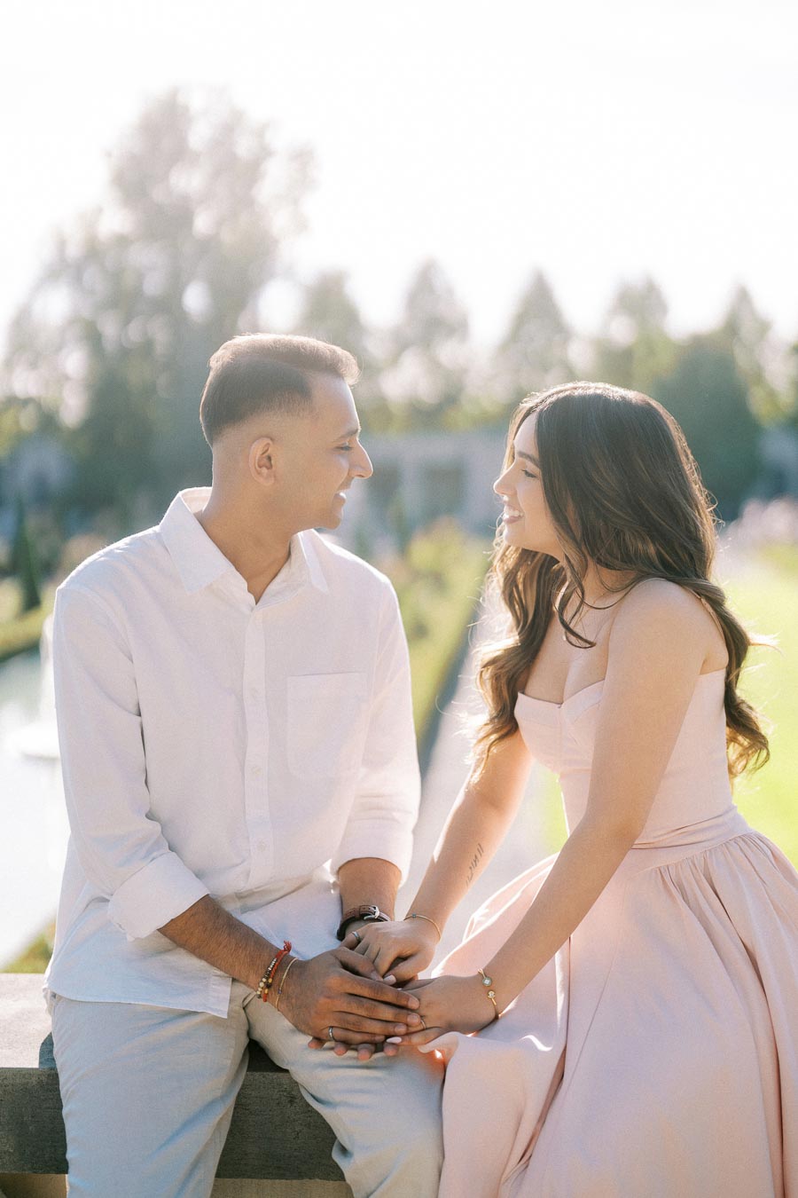 A couple sitting outdoors, holding hands and smiling at each other on a sunny day, with a scenic background of greenery and a river.