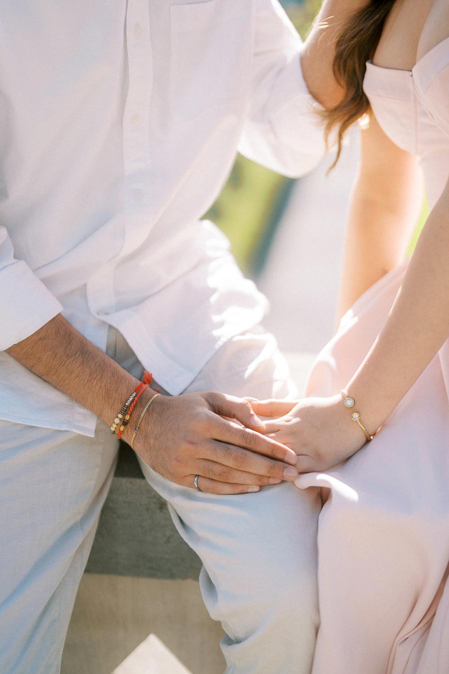 A couple sitting close together wearing light-colored clothing, showcasing hands gently holding each other, with focus on the woman's delicate bracelet and soft pink dress, conveying a sense of intimacy and romance in a sunny outdoor setting.