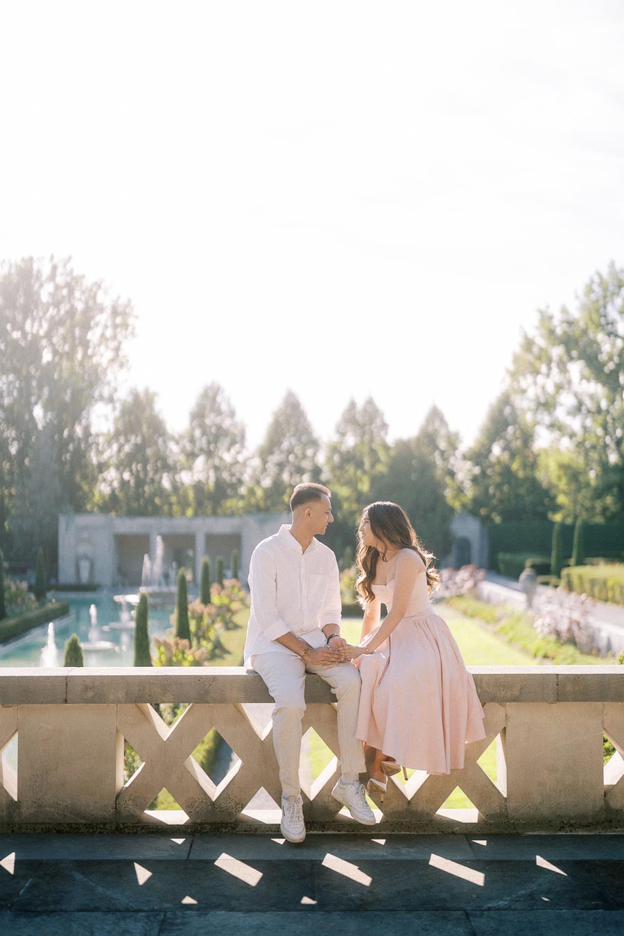 A couple sitting on a stone wall in a sunlit garden, dressed in light pastel colors, with a fountain and manicured hedges in the background.