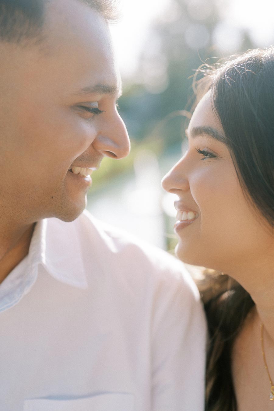A couple smiling at each other closely, surrounded by soft natural light and a blurred outdoor background, capturing a romantic and joyful moment.