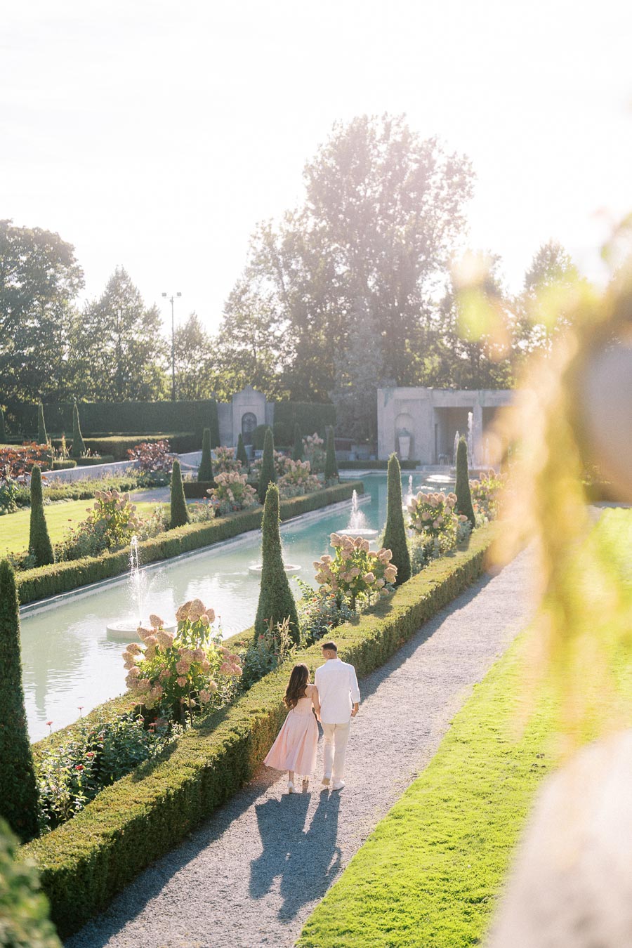 A couple strolling through a picturesque garden with trimmed hedges, a long reflective pool, and vibrant flowers under a bright, sunny sky.