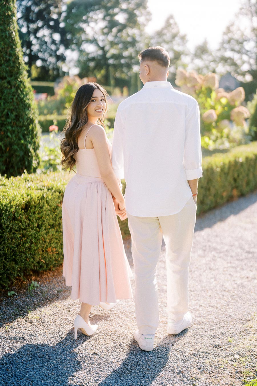 A couple holding hands and walking along a sunny garden path, with lush greenery and flowers in the background. The woman is wearing a pink dress, and the man is in a white shirt and light pants, creating a romantic and serene atmosphere.