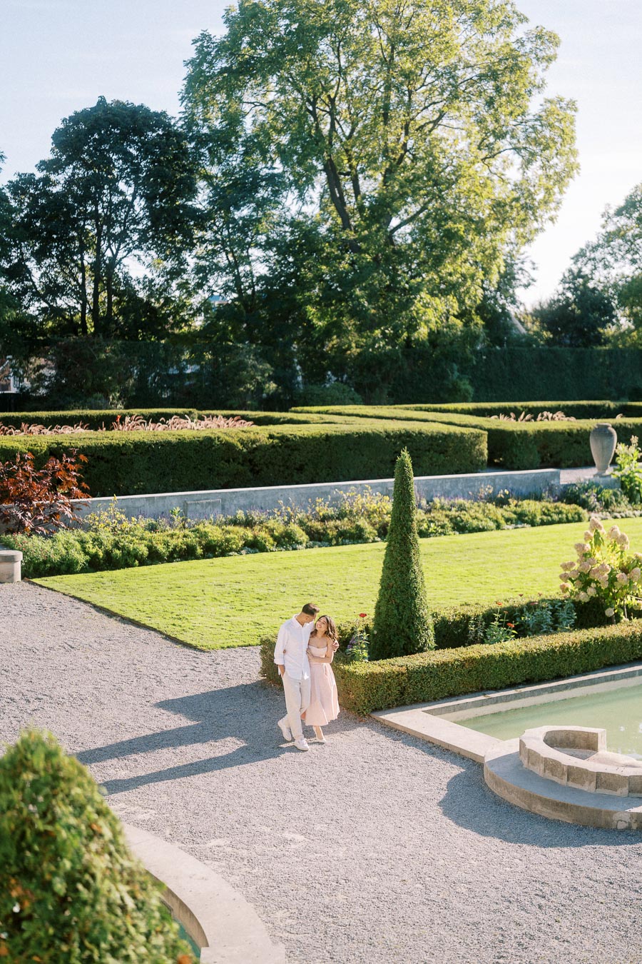 A couple walking hand in hand through a beautifully landscaped garden, surrounded by manicured hedges and lush green trees under a clear blue sky.