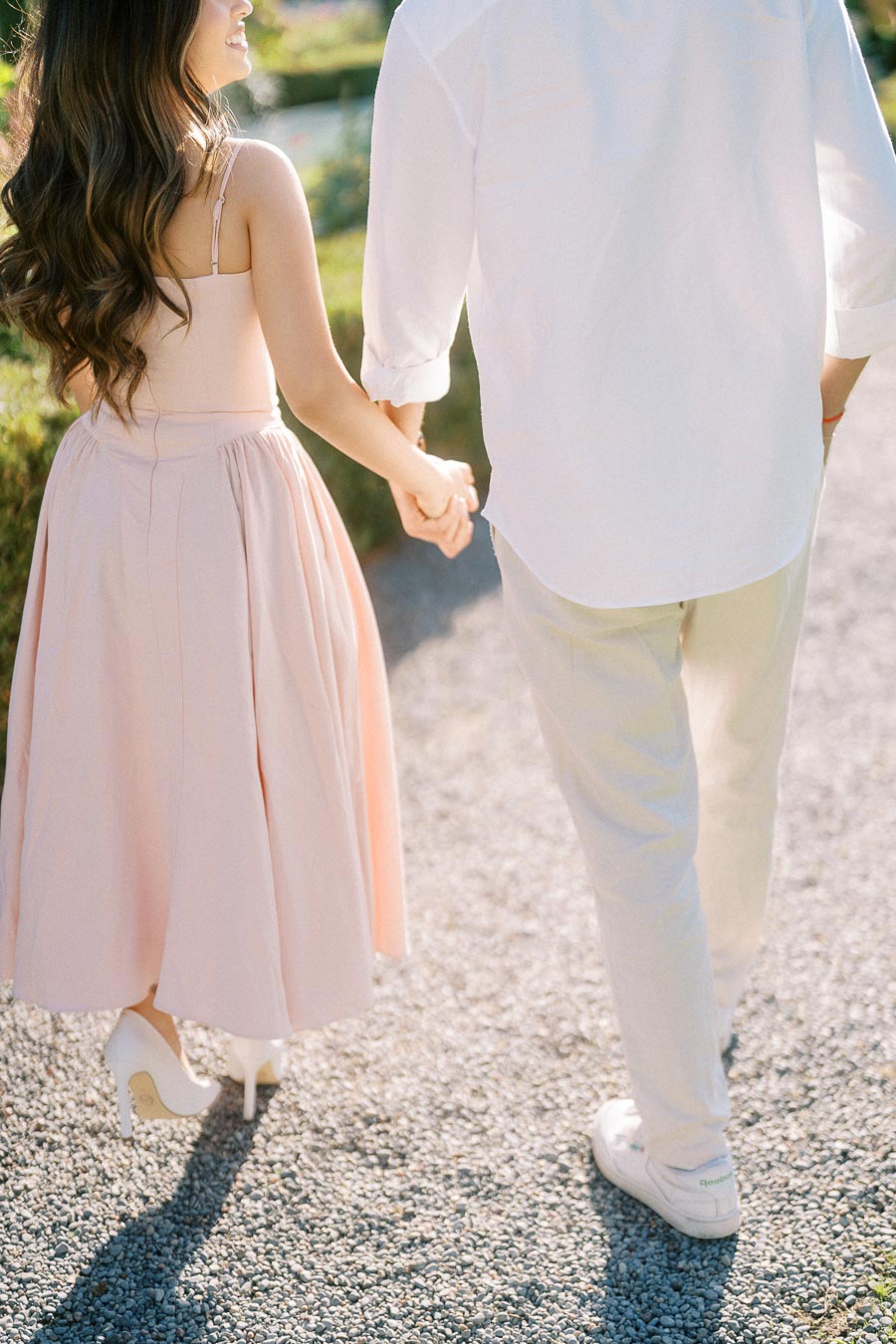 Couple holding hands walking on a sunny day, with woman in a pink dress and man in white shirt and pants, on a gravel pathway in a garden setting.