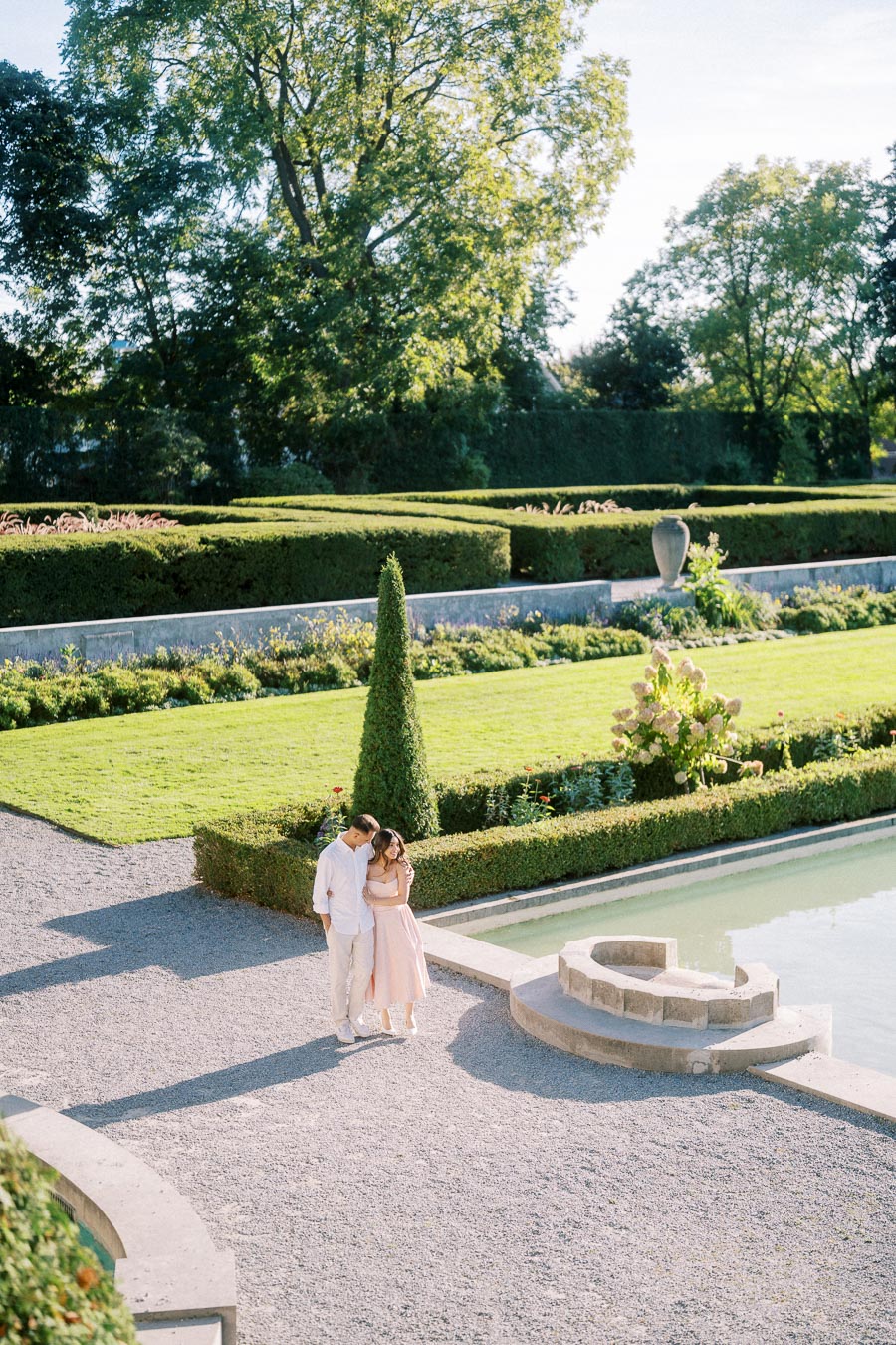 A couple embracing by a serene garden pond, surrounded by meticulously trimmed hedges and lush greenery on a sunny day.