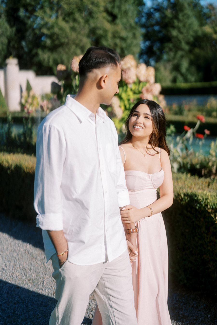 A couple walking hand in hand in a garden setting, with the woman in a pastel pink dress looking up and smiling at the man in a white shirt.