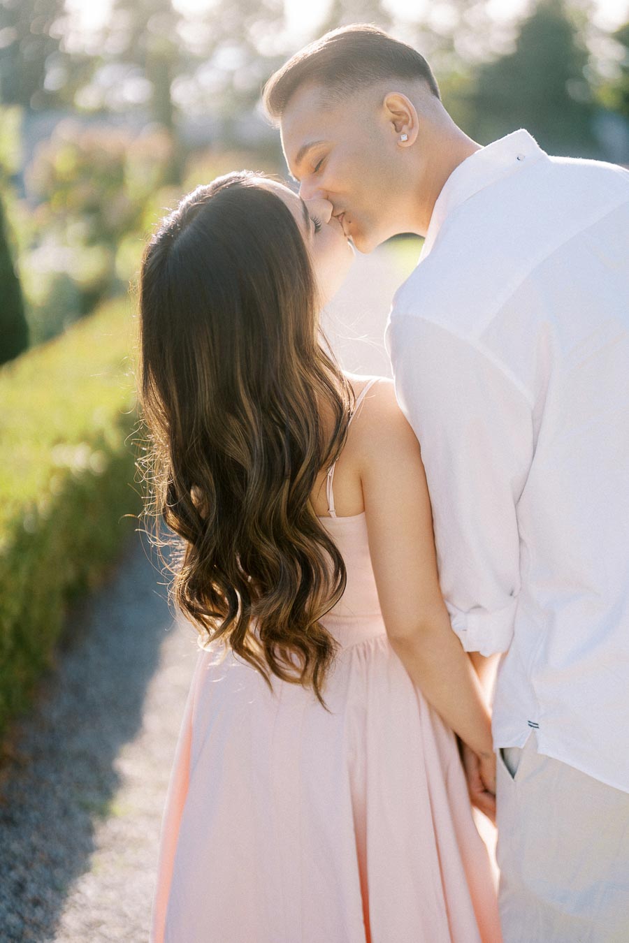 A couple shares a romantic kiss in a sunlit garden path, with lush green hedges and soft sunlight creating a picturesque, serene setting.