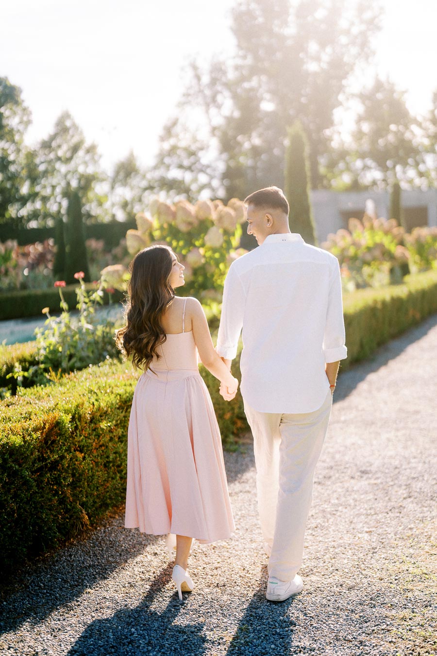 A couple holding hands while walking through a sunlit garden path, surrounded by lush greenery and vibrant flowers, capturing a romantic and serene moment.