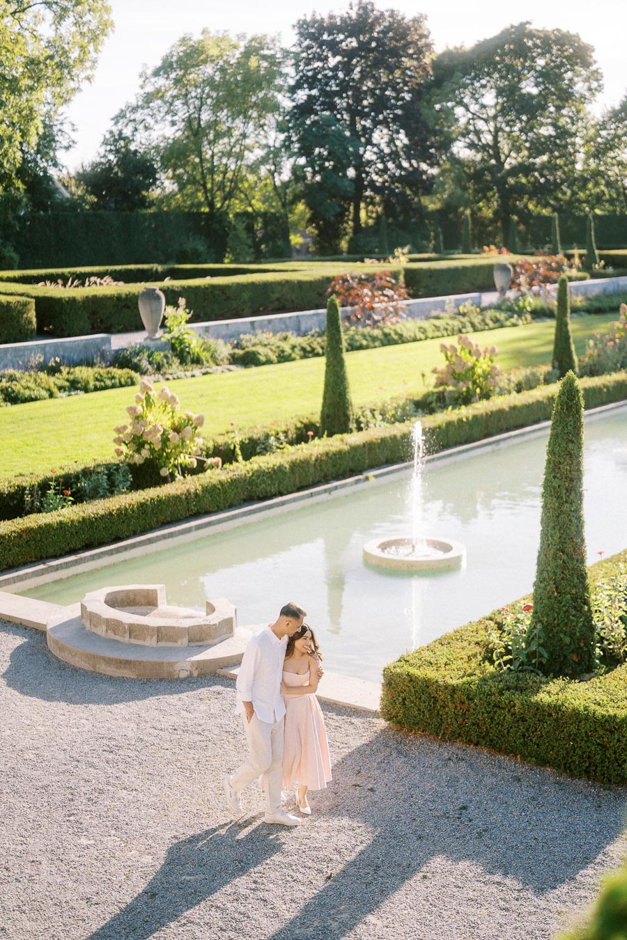 A couple strolling through a picturesque garden with manicured hedges and a fountain pool on a sunny day, surrounded by lush greenery and vibrant flowers, creating a romantic and serene atmosphere.
