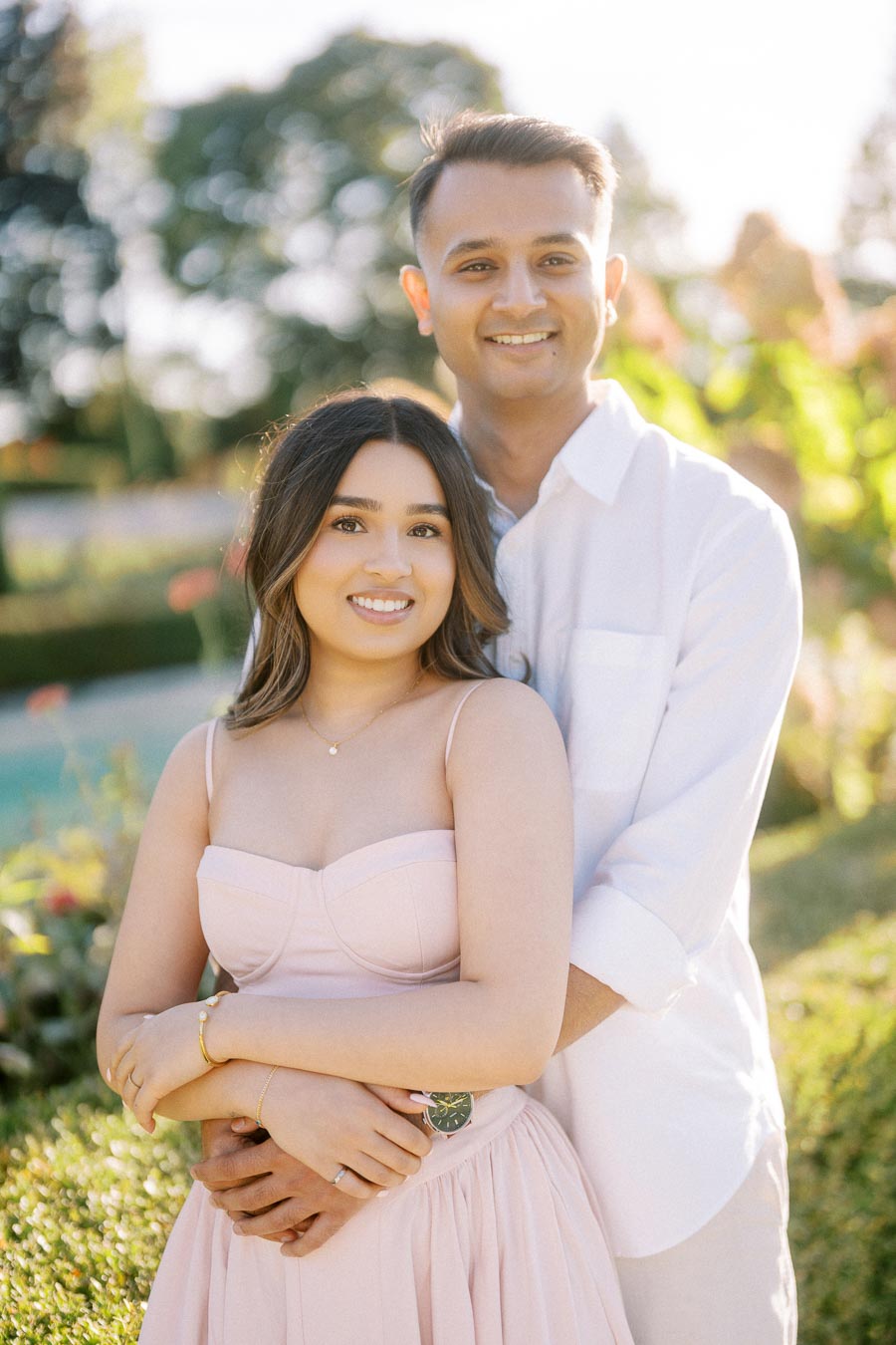 Couple embracing and smiling in a sunlit garden, both wearing white and pink outfits, showcasing a romantic outdoor setting.