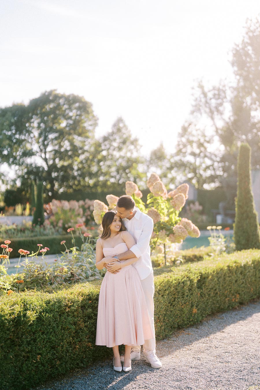 A couple in elegant attire embracing in a lush garden surrounded by blooming flowers and greenery, under a bright sunny sky.