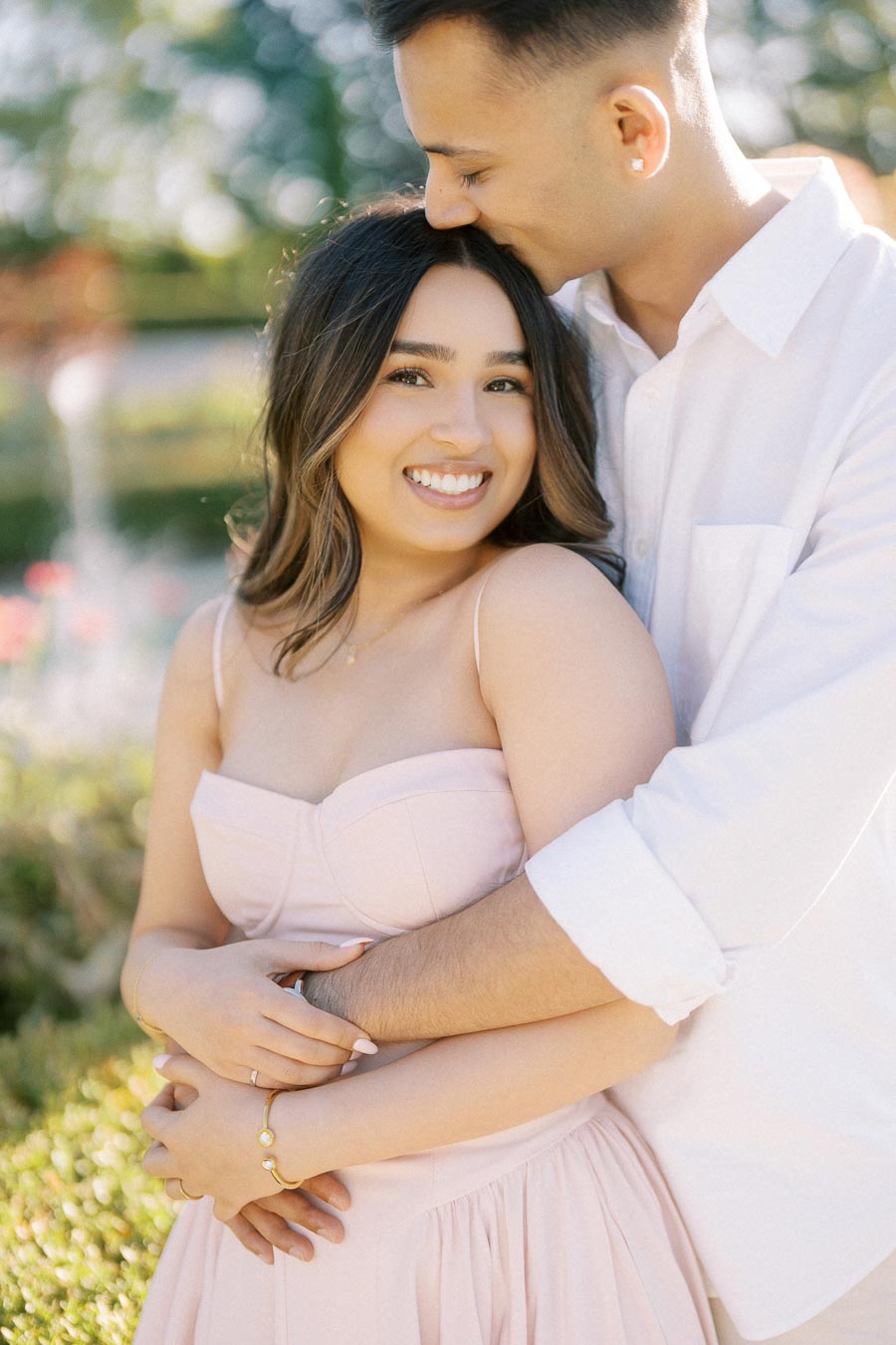 Smiling couple embracing in a sunny garden, with the woman in a light pink dress and the man in a white shirt, showcasing love and happiness.