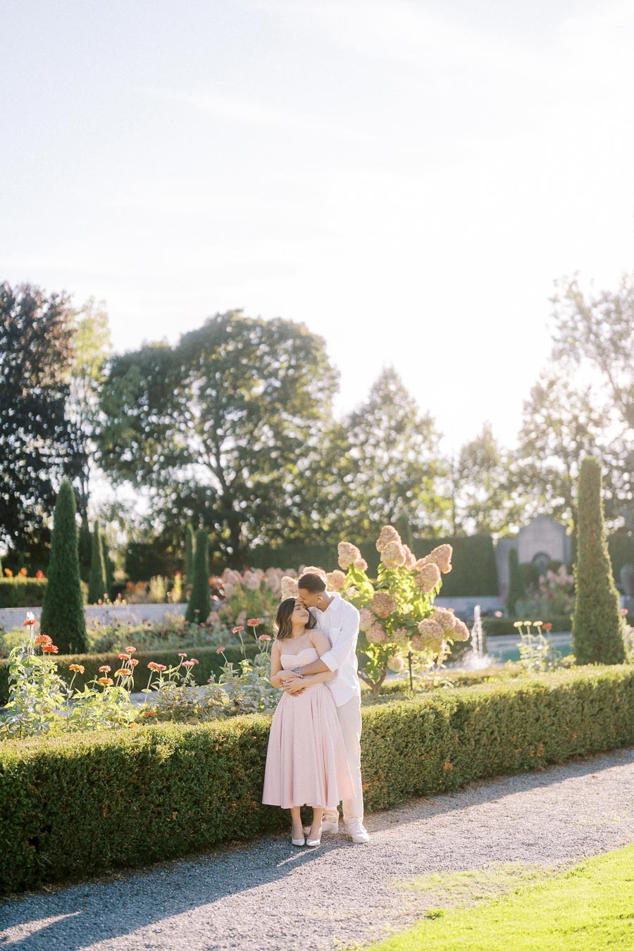 A romantic couple embracing in a beautiful sunlit garden with blooming flowers and tall trees on a clear day.