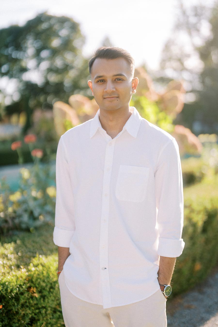 Man in a white shirt standing outdoors in a sunlit garden with blurred greenery in the background.