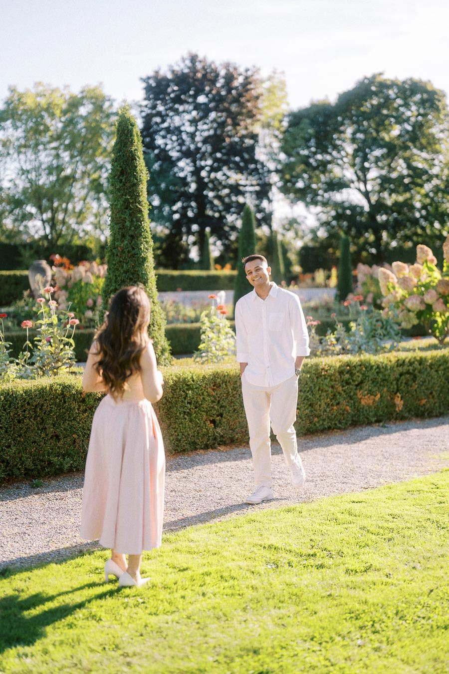 A couple enjoying a sunny day in a beautifully landscaped garden, surrounded by vibrant flowers and manicured hedges. The man is smiling at the woman, capturing a romantic moment.
