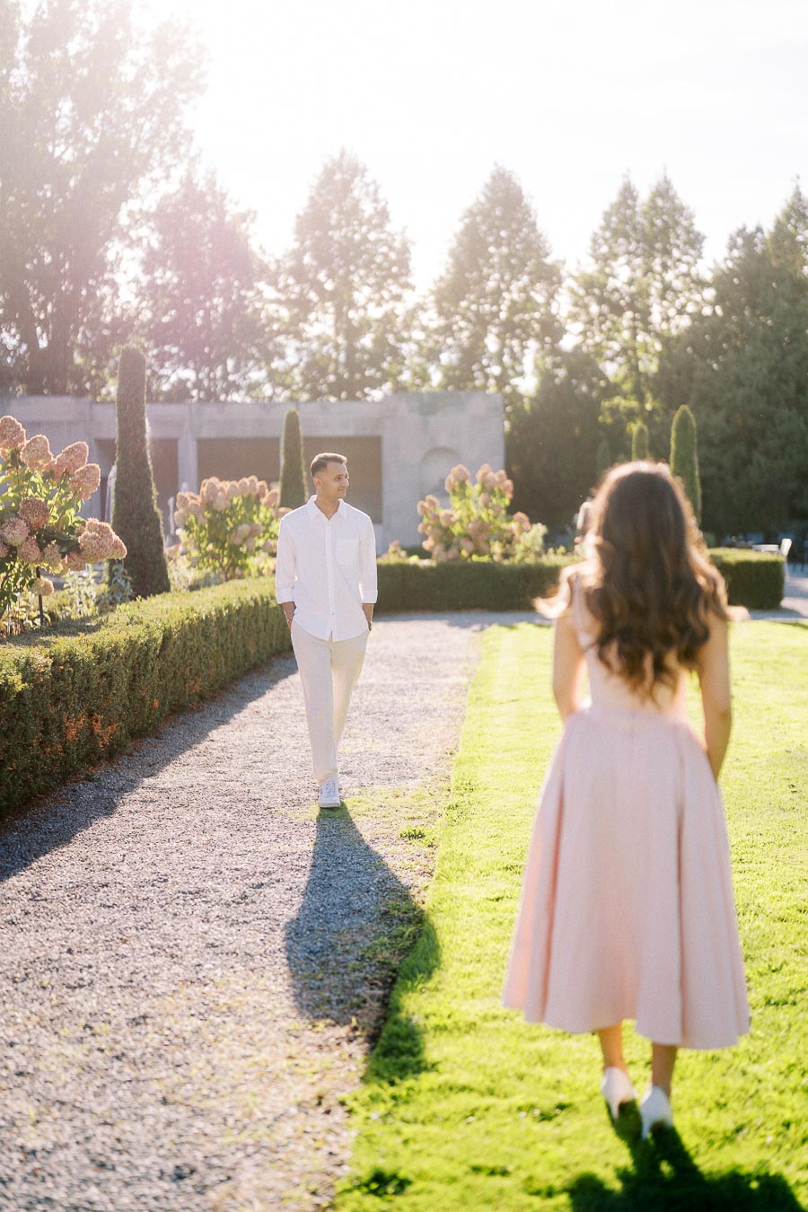 A couple in a sunlit garden, with the man wearing a white shirt and pants walking on a gravel path towards a woman in a pink dress, surrounded by vibrant greenery and flowers in the background.