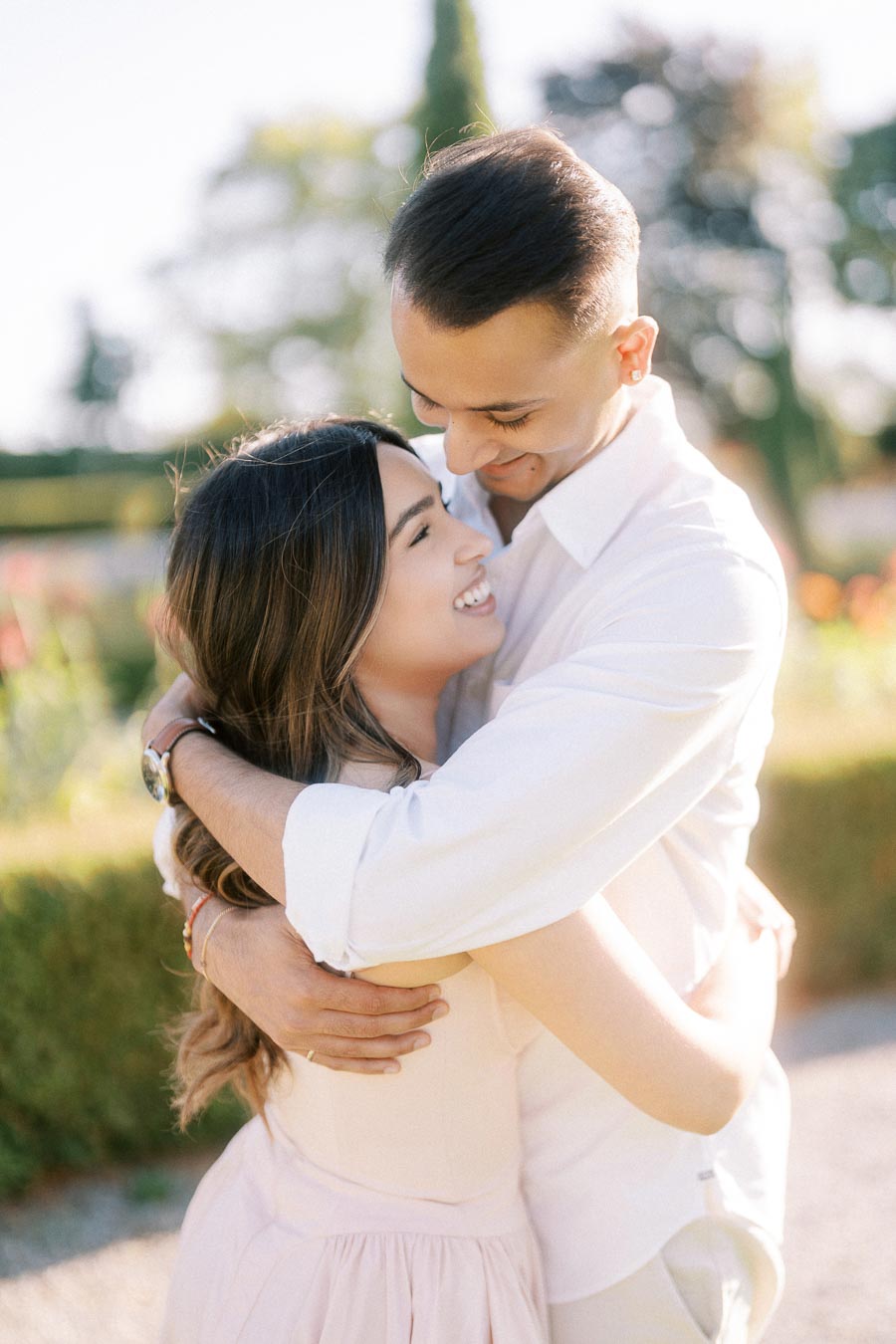 A couple embracing and smiling warmly at each other in a sunlit garden setting, wearing light-colored clothing.