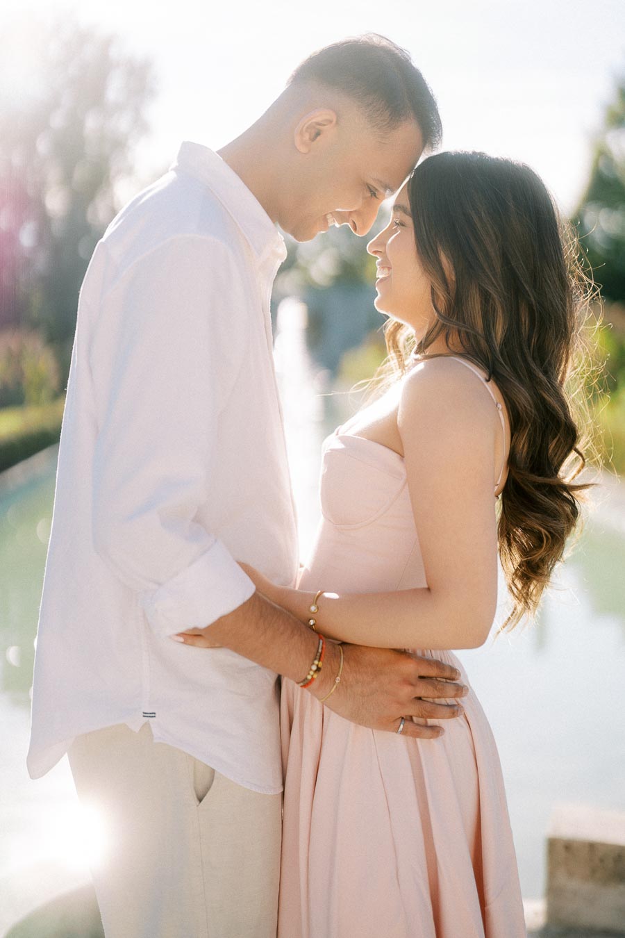 A romantic couple embracing and smiling at each other, with a sunlit garden background. The man is in a white shirt, and the woman is wearing a light pink dress, conveying love and happiness in a picturesque outdoor setting.