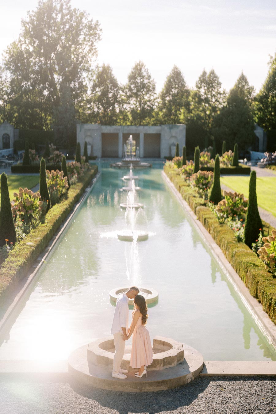 Romantic couple holding hands by a scenic fountain in a beautifully manicured garden, surrounded by lush greenery and topiary trees, under a bright blue sky.