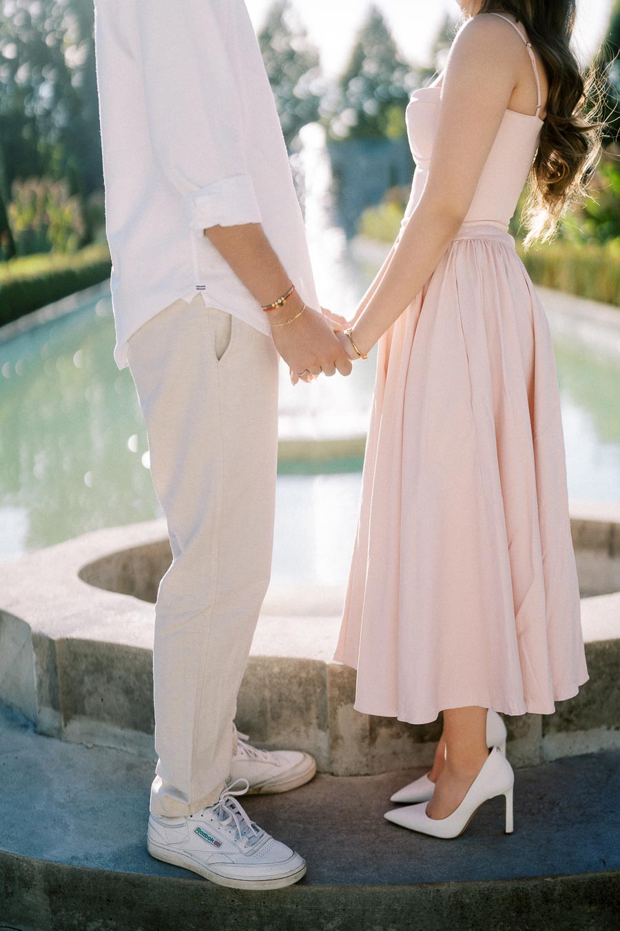 A couple holding hands at an outdoor setting, with a woman in a pink dress and white heels, and a man in casual white attire and sneakers, standing near a fountain on a sunny day.