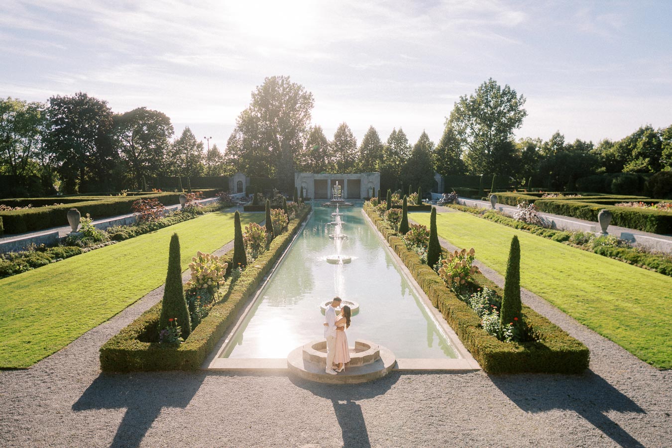 Romantic couple embracing by a serene garden reflecting pool with symmetrical landscaping, vibrant green lawns, and elegant fountains under a clear blue sky.