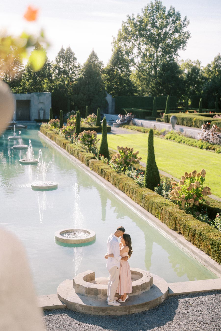 A romantic couple embracing by a picturesque fountain in a lush garden setting, with manicured hedges and trees under a clear blue sky.