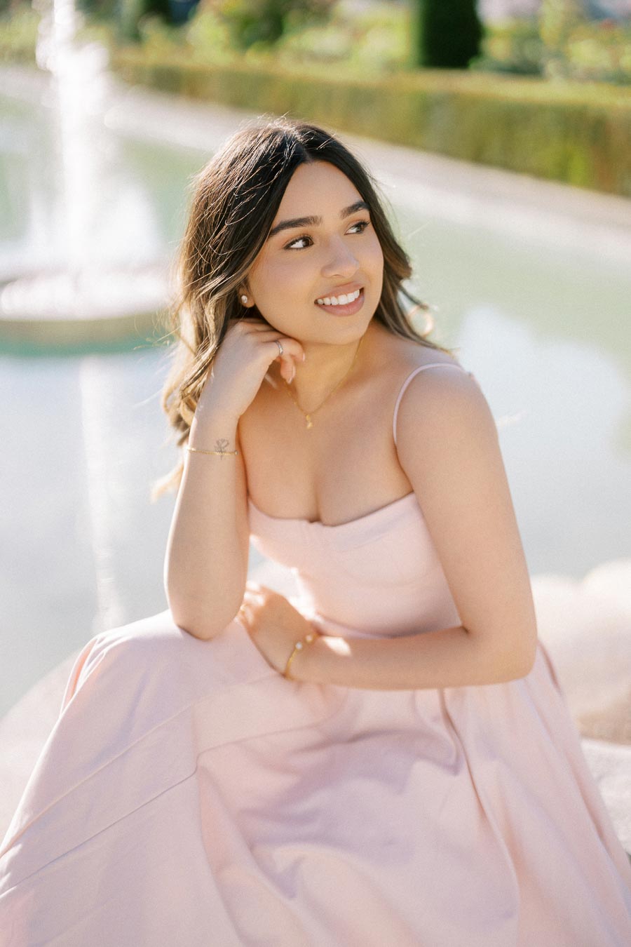 Young woman in a light pink dress sitting by a fountain, looking away with a gentle smile, outdoors on a sunny day.