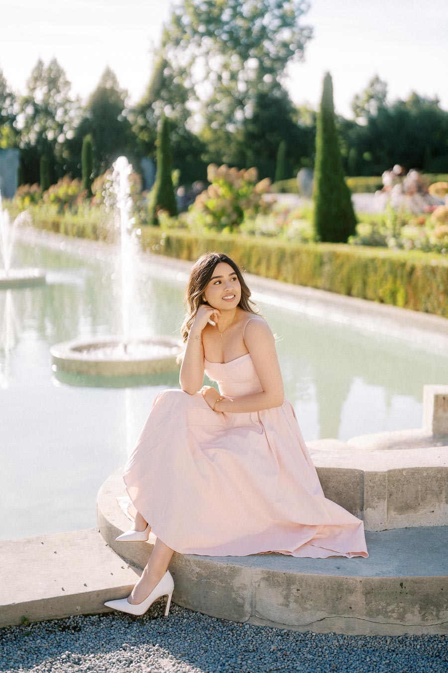A woman in a pink dress sits gracefully by a fountain in a botanical garden on a sunny day, surrounded by lush greenery and trees.
