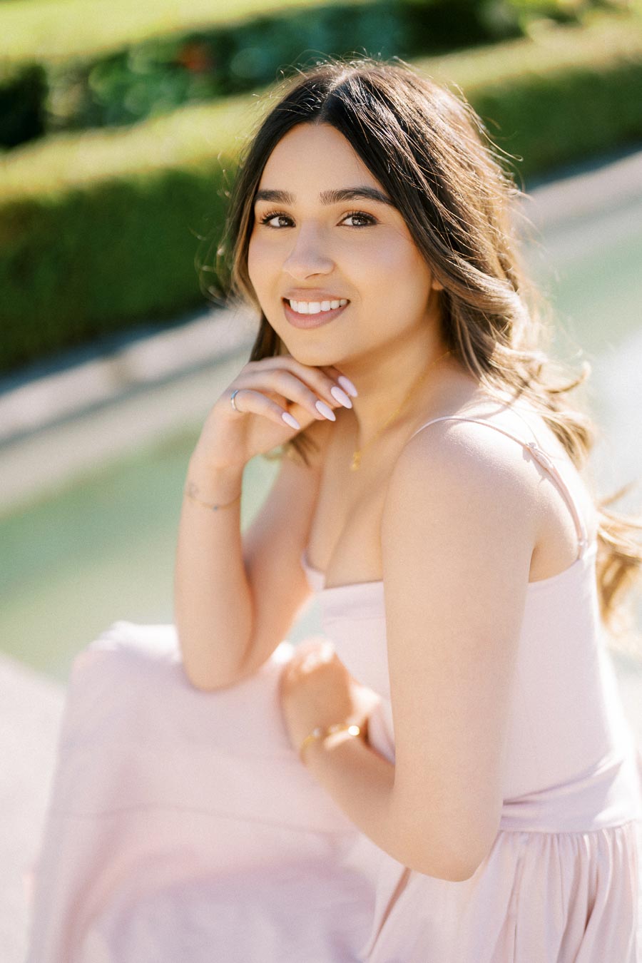 Young woman in a light pink dress smiling outdoors, sitting by a serene pool with greenery in the background.