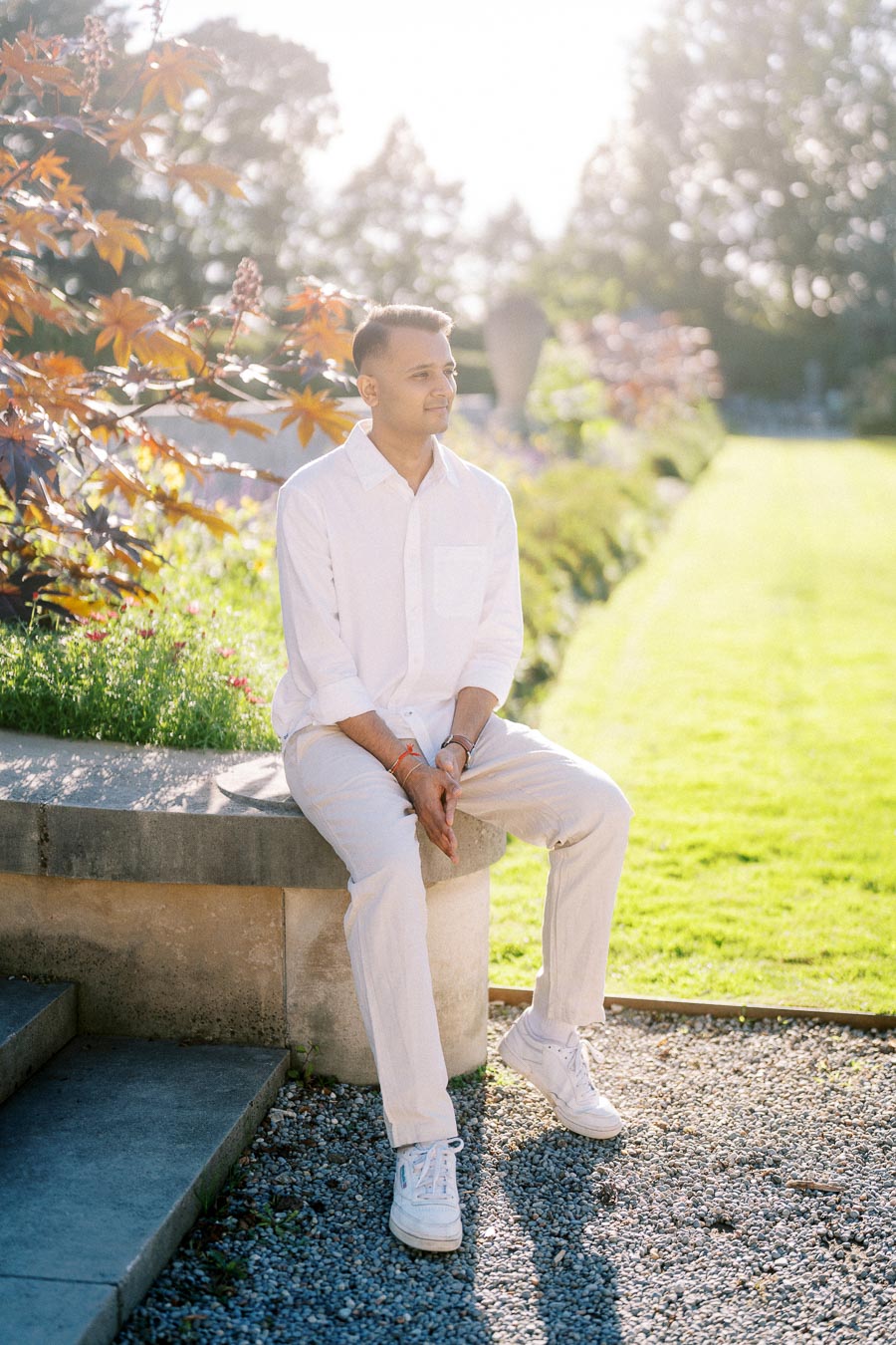 A person in a white outfit sitting on a stone ledge in a sunlit garden, surrounded by greenery and autumn-colored leaves, enjoying a tranquil moment in nature.