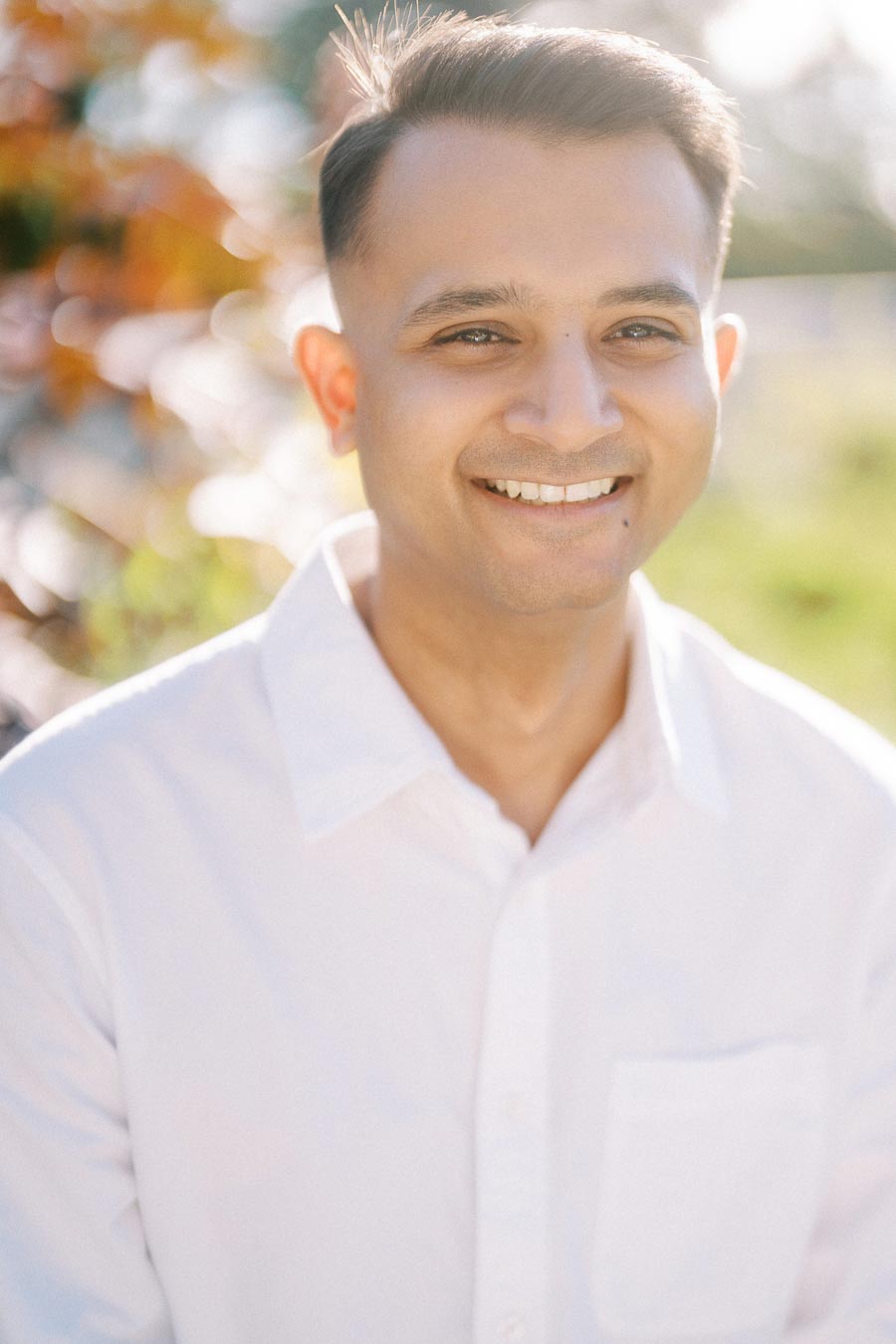 Smiling person outdoors in sunlight wearing a white collared shirt.
