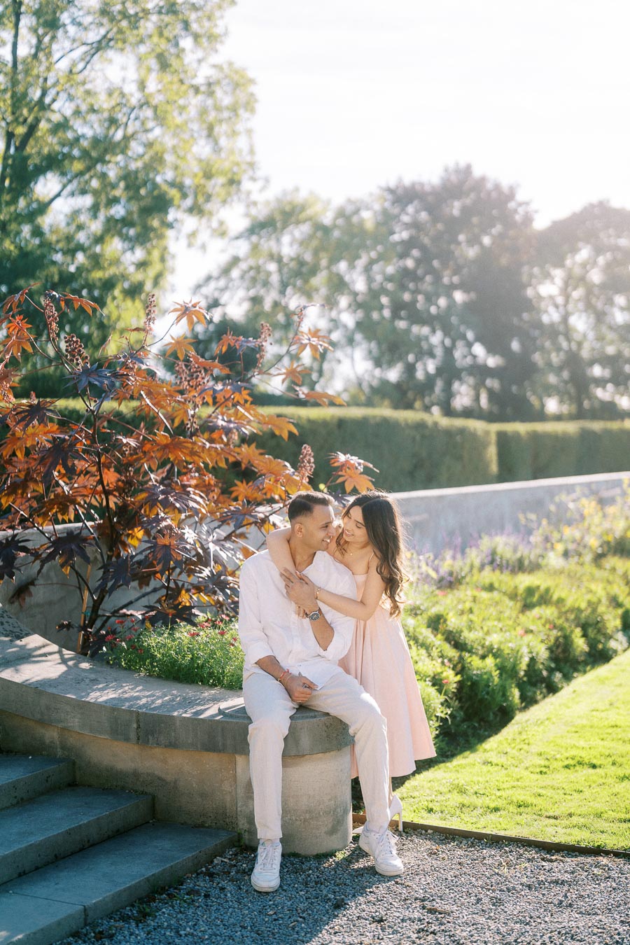 A couple enjoying a romantic moment in a sunlit garden, surrounded by vibrant greenery and ornamental plants, with a serene atmosphere and clear blue sky in the background.