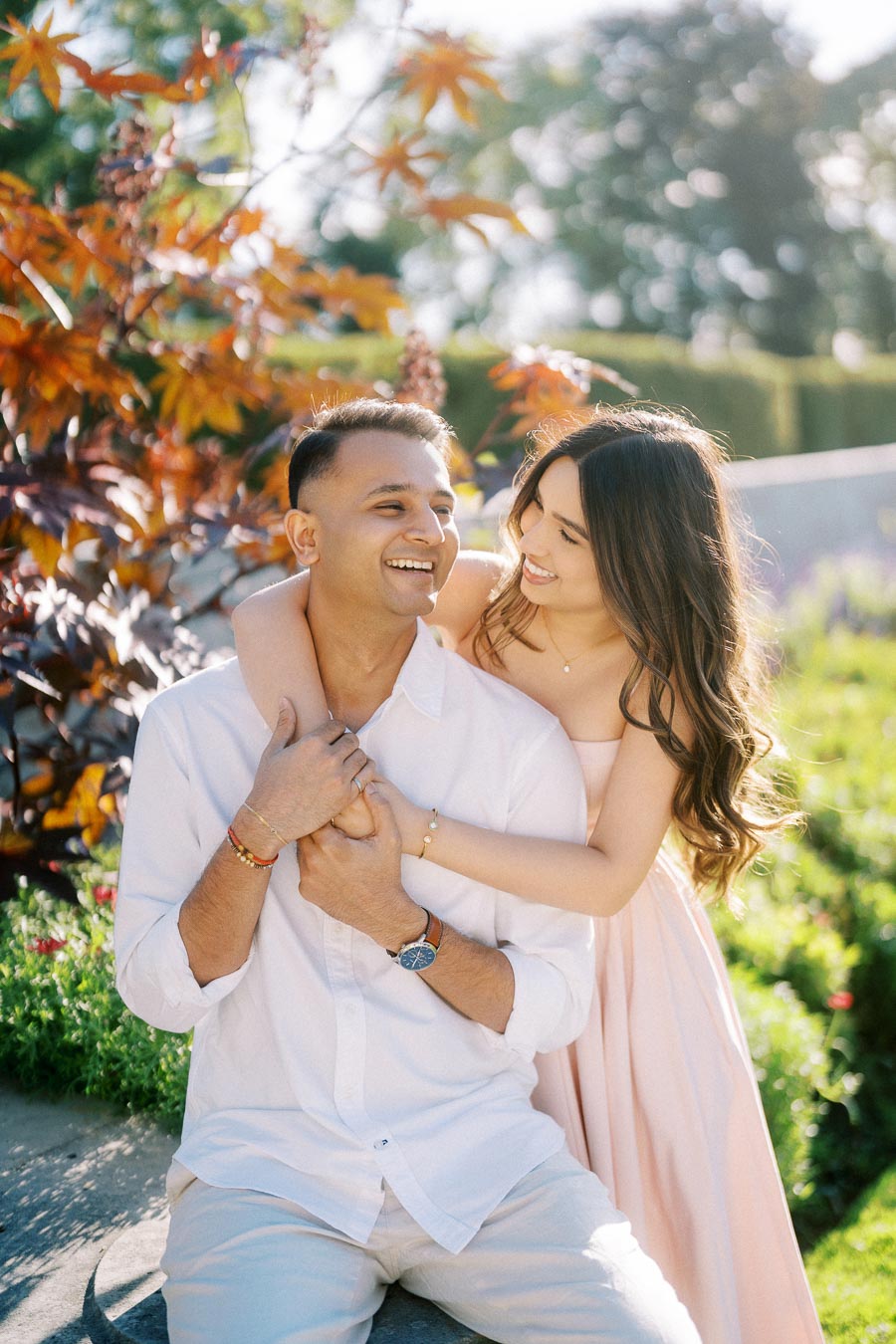 A happy couple embraces in a sunlit garden, surrounded by colorful autumn leaves and greenery, sharing a joyful moment.