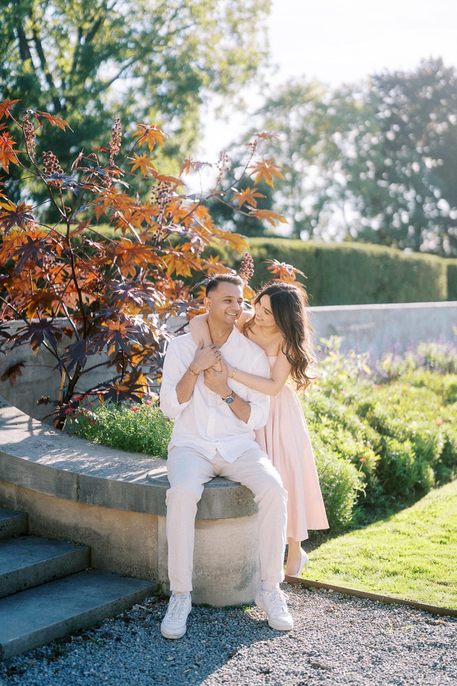 A couple enjoying a sunny day in a garden, sitting on a stone wall surrounded by vibrant green plants and trees, smiling and embracing each other.