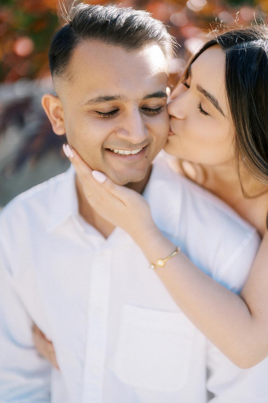 A woman gently kisses a smiling man on the cheek, creating a romantic and affectionate scene. The man wears a white shirt, and the background features soft, blurred autumn foliage, adding warmth to the image.