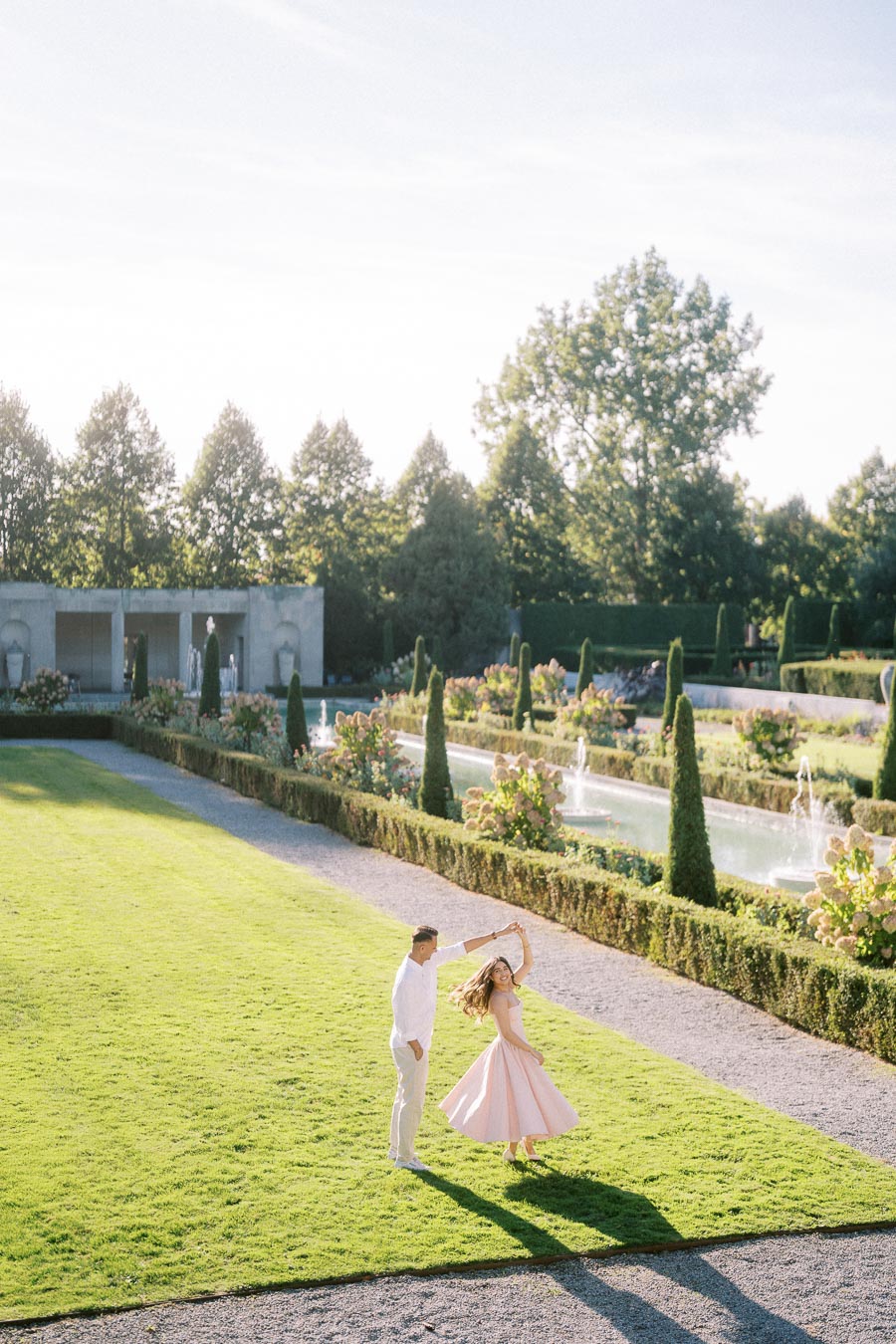 A couple dancing on a lush green lawn in a picturesque garden with topiary and fountains under a clear blue sky.