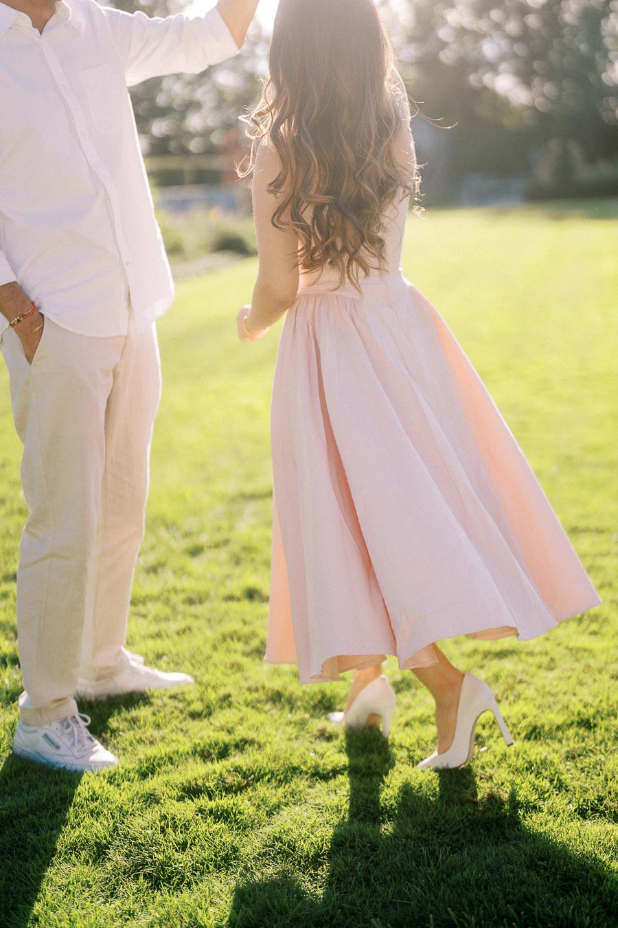 A couple dancing on a sunlit lawn, wearing light-colored clothing; woman in a blush pink skirt and heels, man in white shirt and sneakers, enjoying a romantic moment.