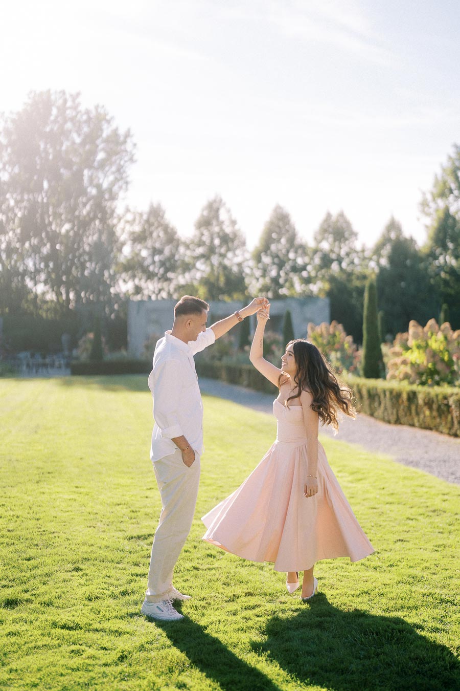 A couple dancing gracefully on a sunlit garden lawn with trees and hedges in the background, featuring the woman in a flowing peach dress and the man in a white shirt and beige pants.