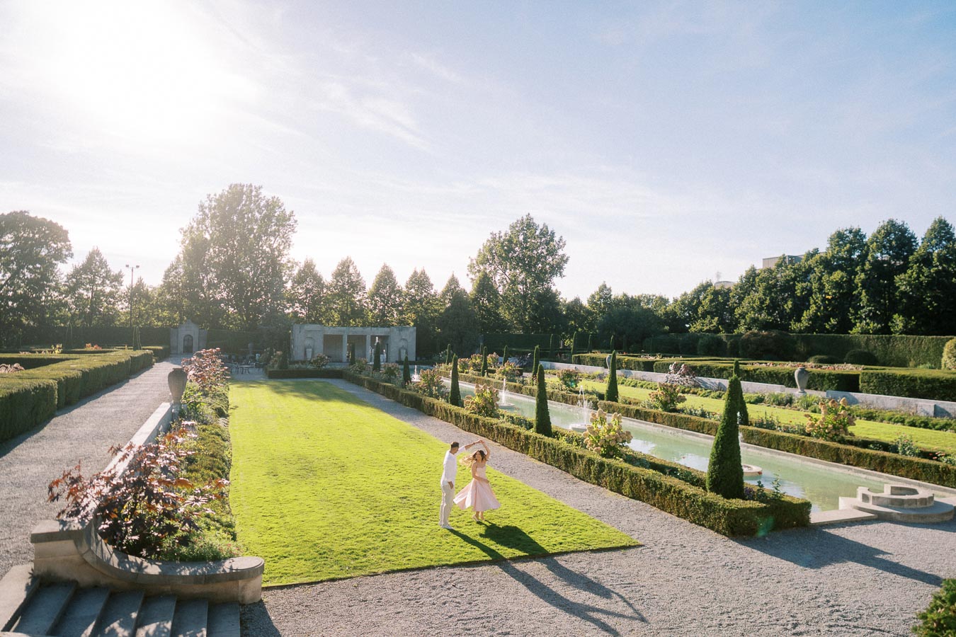 A couple dancing on a lush green lawn in a sunlit garden with symmetrical paths, hedges, and a reflecting pool under a clear blue sky.