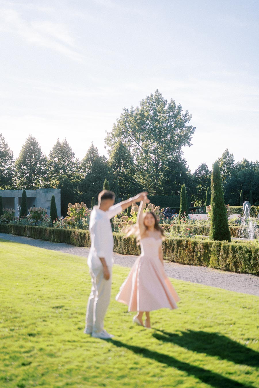 A couple dancing joyfully on a sunlit garden lawn, surrounded by lush greenery and fountains in the background, under a clear blue sky.