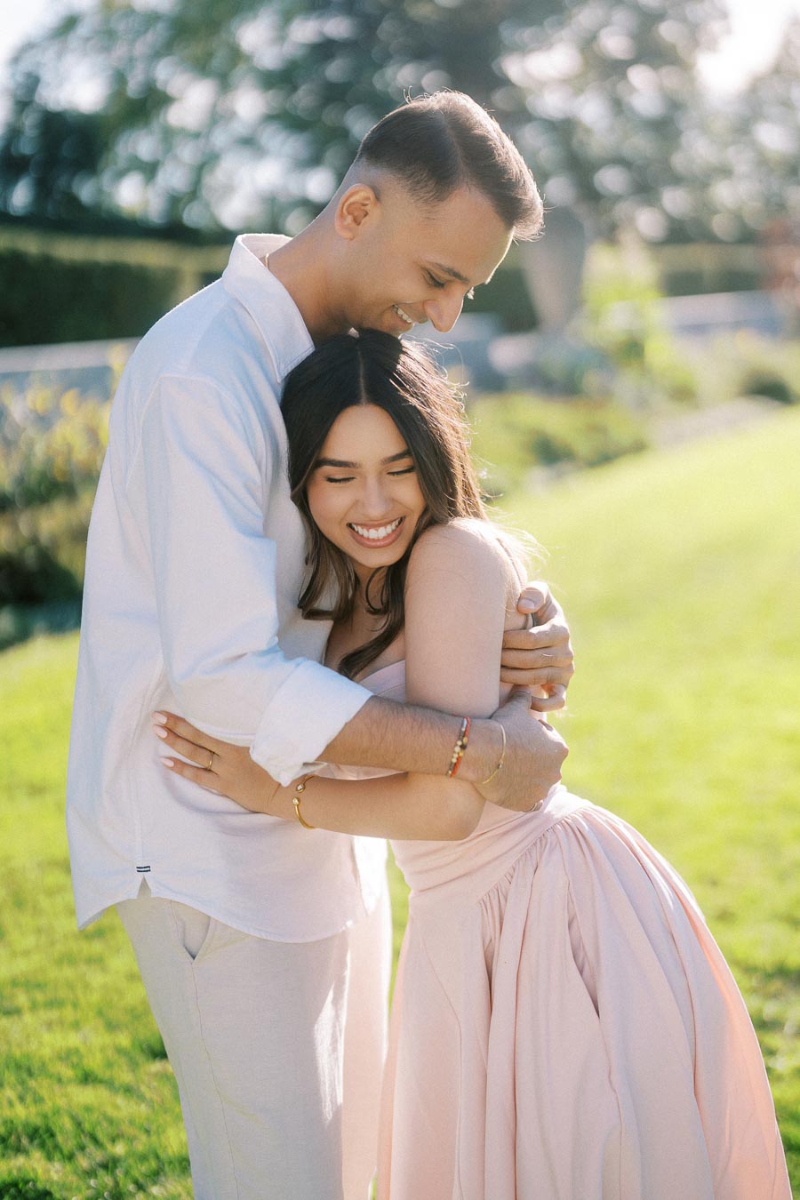 A joyful couple embraces in a sunlit garden, with the man wearing a light shirt and the woman in a pink dress, both smiling warmly, surrounded by lush greenery.
