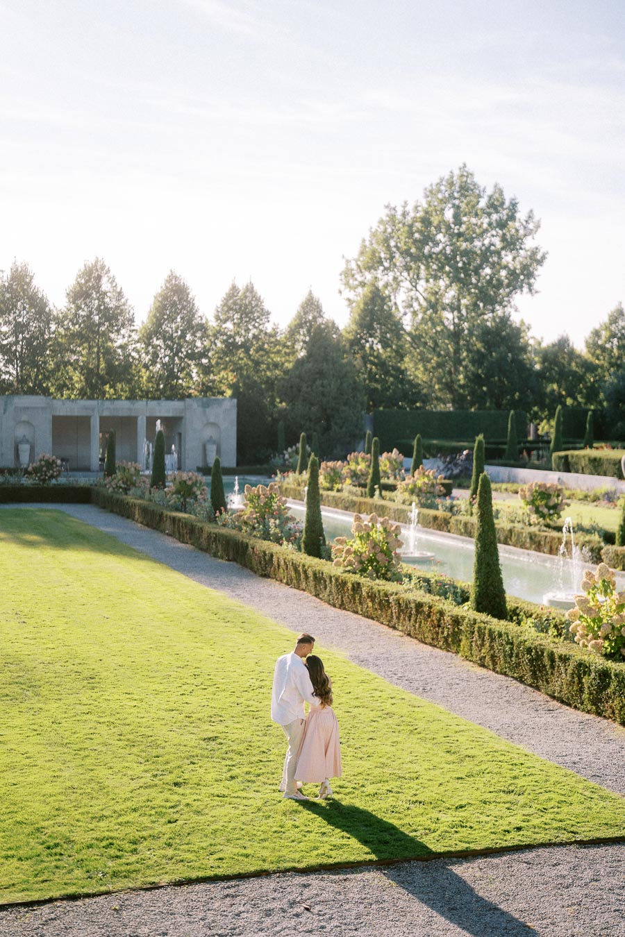 A couple walking hand in hand across a sunlit garden with neatly trimmed hedges and fountains, surrounded by lush greenery and a classic pavilion in the background.